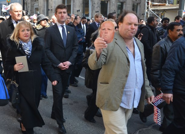 People walking in a procession including a man holding a Rob Ford mask