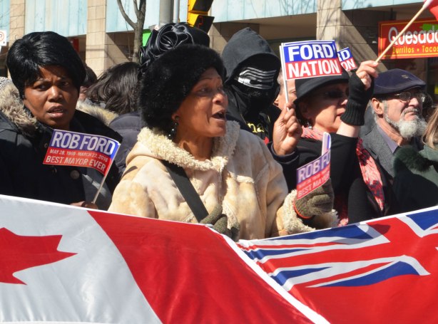 a man in a black and white mask stands behind some women waving ford nation flags. 