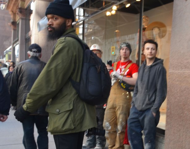 A black man with beard and moustache turns to look back, three young men in work clothes stand against the storefront beside and behind him. 