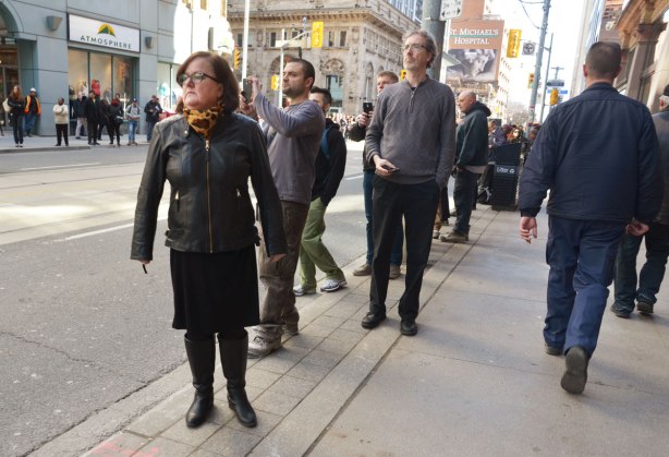 a small group of people wait on the sidewalk, watching down the street, one man with a camera in hand. 