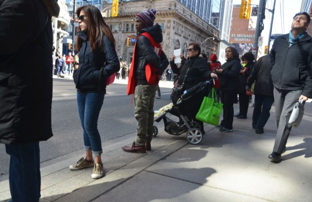 a small group of people wait on the sidewalk, watching down the street, one woman with a camera in hand.