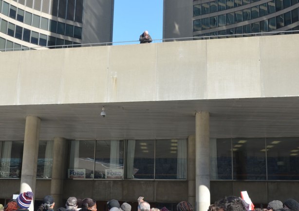 A lone cameraman stands on the upper level at City Hall outside, taking pictures of the people below. 