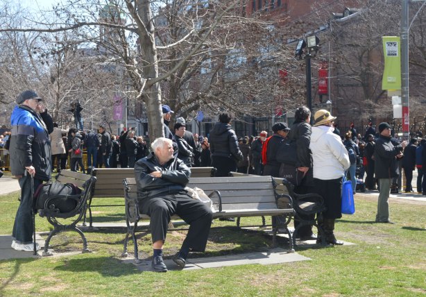 An older man sits on a bench in front of St. James cathedral while other people stand around, watching the procession for Rob Ford's funeral