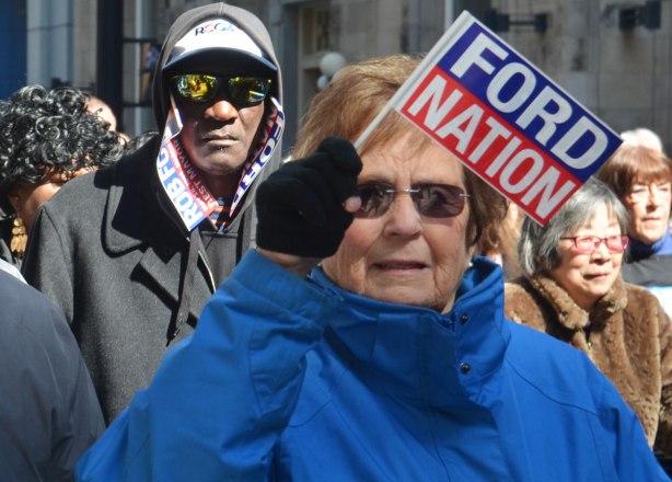An older woman waves a little Ford Nation flag while the man behind her has used Ford Nation signs in lieu of a scarf. He is wearing reflective sunglasses too. 