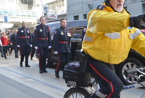 A police man in a yellow jacket and on a bike is trying to get the crowd to stand back as he rides beside a hearse with police guard as it drives down the street. 