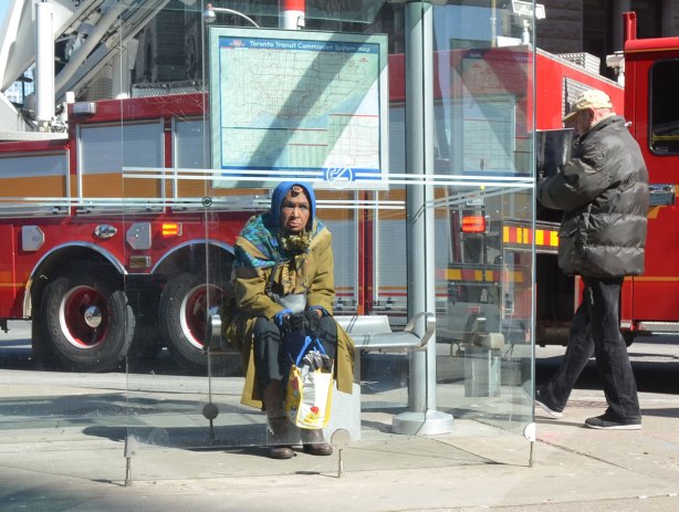 An older woman sits on a bench inside a TTC bus shelter. A fire truck is behind her. 