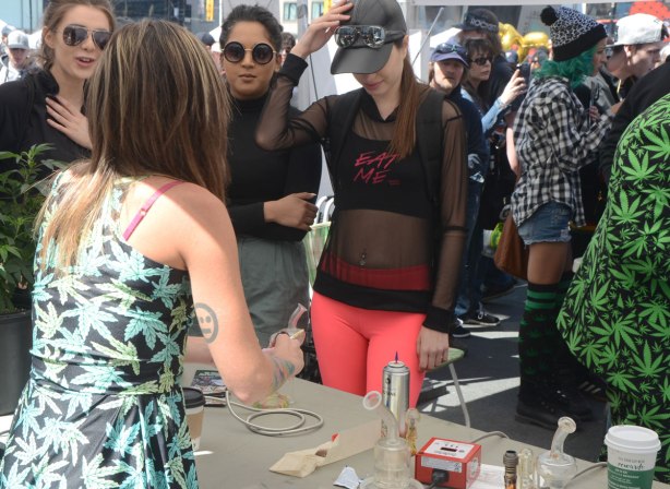 People at Yonge Dundas Square in Toronto celebrating 420 day - three young women in sunglasses are talking to a vendor at the event. One is wearing bright orangish pink tight pants and a black top with the words eat me on it. 