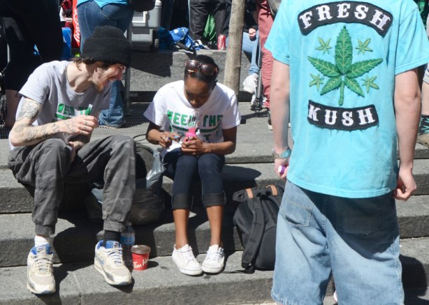 People at Yonge Dundas Square in Toronto celebrating 420 day - two people sitting together and talking, a man's back is to the camera and on the back of his tshirt are the words fresh kush and a picture of a cannabis leaf 
