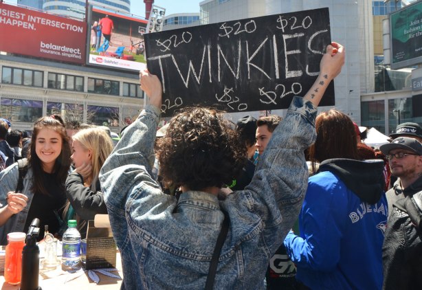 People at Yonge Dundas Square in Toronto celebrating 420 day - a young woman in a jean jacket is holding up a black sign advertising twinkies for sale. 