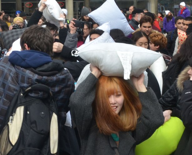 an asian woman with her hair dyed auburn has a pillow over her head as she emerges from a crowd at a pillow fight