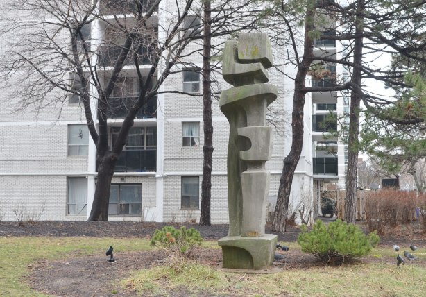tall columnar sculpture somewhat resembling a totem pole, all in grey, beside some trees in front of an apartment building. 