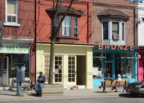 Looking across the street at a man sitting on the edge of a concrete planter for a tree as well as a multicoloured striped bench with two women sitting on it. They are in front of two storey brick buildings with stores on the bottom level and apartments on the top. One of the stores is Bronze.