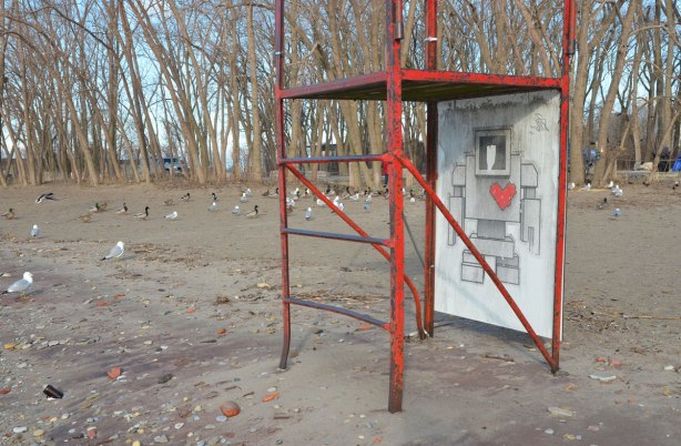 an empty red metal frame lifeguard chair on the beach in early spring, no leaves on the trees. Cherry Beach, Lake Ontario. There is a large lovebot wheatpaste on the lifeguard chair. 