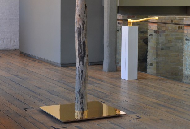 picture taken inside an art gallery - a tree trunk stands in the middle of a gold toned mirror, in the background, a gold plated (or gold colured) axe with its blade in the top of a white rectanguar column 