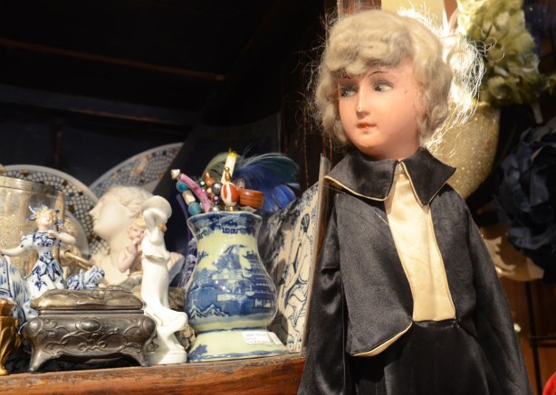 A small doll with a very lifelike face is looking towards a shelf filled with small ceramic and porcelain items such as vases and figurines