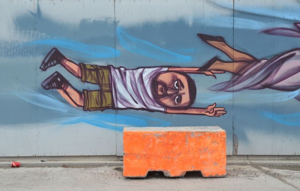 the end of a mural, a man is sideways, arm above his head, painted on a mural, a large orange concrete block is in front of the wall, an empty Tim Hortons cup (Timmies cup) is on the ground. 