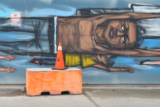 Two people painted flying sideways on a mural, a brown man with no shirt on and a much smaller person below him with a yellow T-shirt. An orange concrete block is on the sidewalk in front of the mural along with an orange and white traffic cone