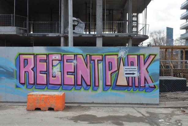 the words Regent Park are written in pink paint on a light blue background, painted on wooden hoardings around a construction site.