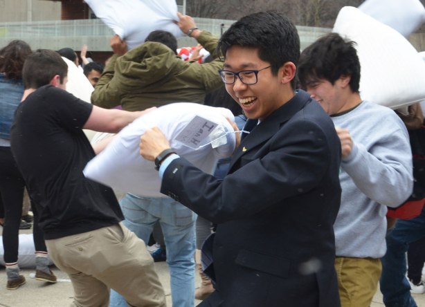 asian young man in suit and tie and glasses is laughing and smiling as he swings a pillow in a pillow fight outdoors with many people