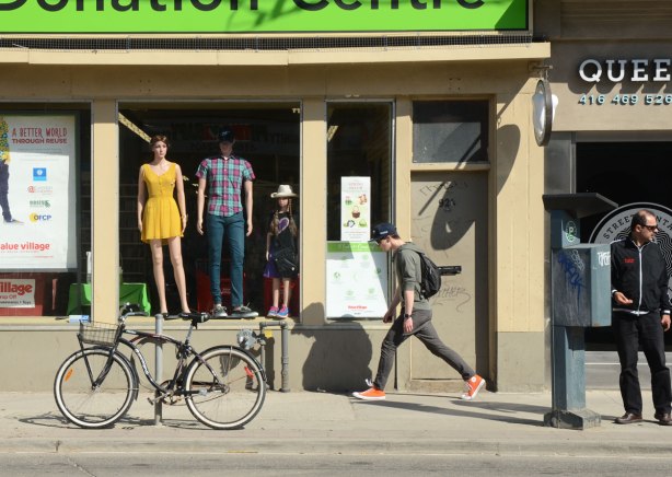 A young man walks past the Value Village Donation Center that has a large window with 3 mannequins in it. A bike is parked in front.
