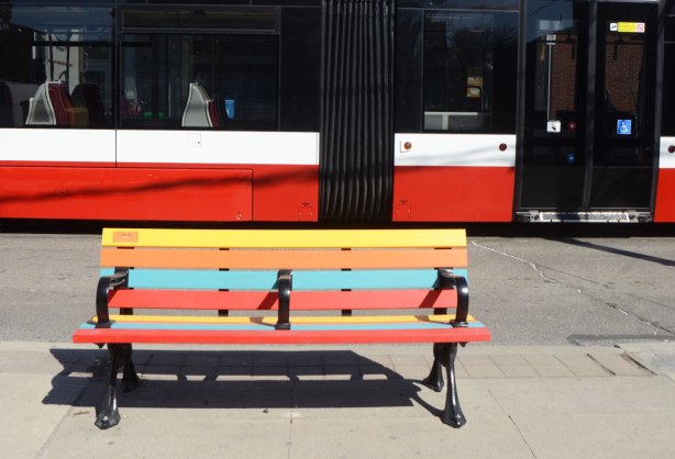 The side of a new TTC streetcar behind a striped bench on a sidewalk