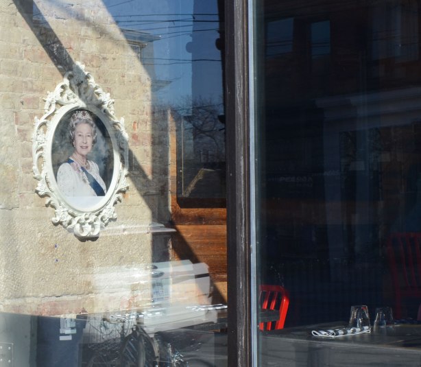 A picture of Queen Elizabeth hangs on a wall in a cafe, seen through the window with reflections of the sky. There are red chairs in the cafe