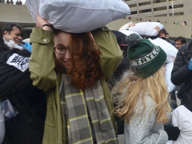 people in the midst of a large pillow fight at Nathan Phillips square in celebration of international pillow fight day - a woman with long red hair hides under her pillow as she walks through a crowd