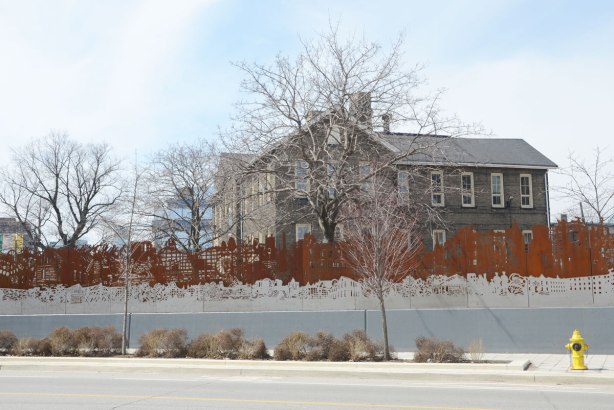 public art on Sumach St., rusted metal and shiny metal fence with cut outs, looking across the street at it, with older stone building behind it. 