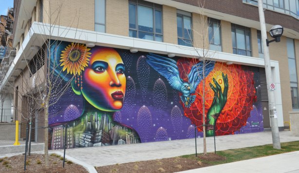 mural painted by Shalak Attack and Bruno Smoky in Regent park Toronto, showing a brightly coloured woman;s face, with a flower in her hair and her body (from the shoulders up) made of buildings in a jumble as well as a blue owl (large bird) in flight, passing in front of the sun that is represented by circles of orange and red radiating out from the bright yellow center. A green hand is reaching up towards the sun 