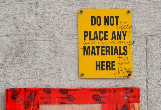A yellow sign in an alley that says "So not place any materials here" on which someone has written in black sharpie