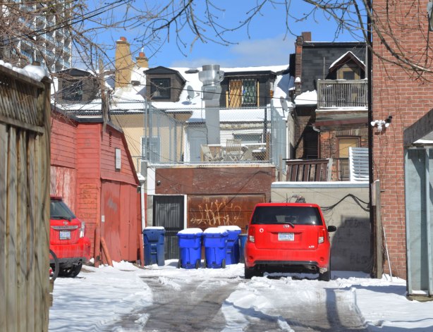 backyard, entrance off alley, little red car parked on snow covered parking lot. blue sky, backs of two storey houses