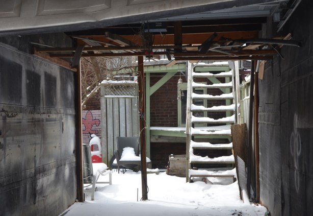 looking through a backless garage where the garage door is open. The garage is empty. you can see the backyard, covered in snow as well as snow covered steps leading up the second storey