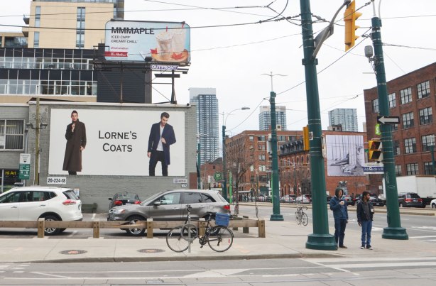 view from the NE corner of Spadina and Richmond streets. a small parking lot is directly across the street, two large billboards loom over the lot. Once is for Lorne's coats showing two people wearing coats. The other is for Maple flavoured iced capp at Tim Hortons.