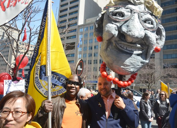 photographs taken at a rally and protest in support of a $15 minimum wage, The Fight for 15 and fairness - two men standing together, one with a United Steelworkers flag and the other with a large effigy of Kathleen Wynne's head held above his own head 