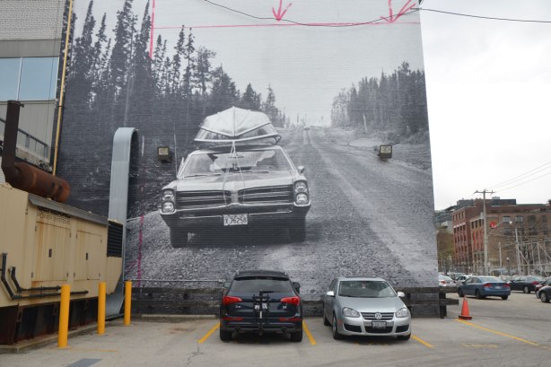 A very large black and white photograph from the 1960's of a car on a two lane road with pine trees on either side. There is a boat tied to the roof of the car. The photo is on the side of the Globe and Mail building with cars parked around it.