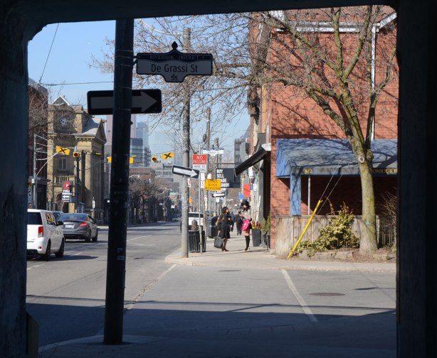 Photo taken from under a bridge, looking west along Queen St. East, cars on the street, a restaurant on the corner, and some women on the sidewalk.