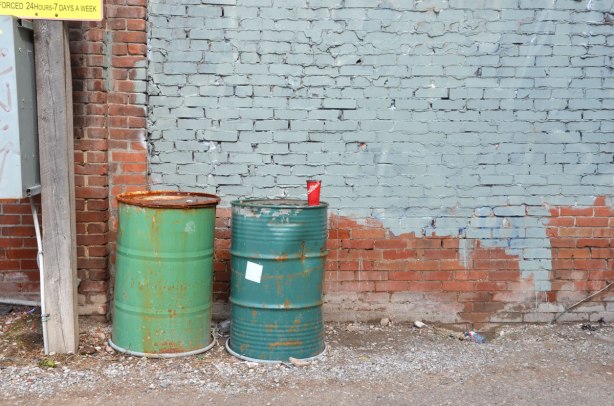 an empty paper coffee cup from Tim Hortons that has been discarded - on top of a dark green barrel that is beside a lighter green barrel, in front of a red brick wall that has been partially painted grey