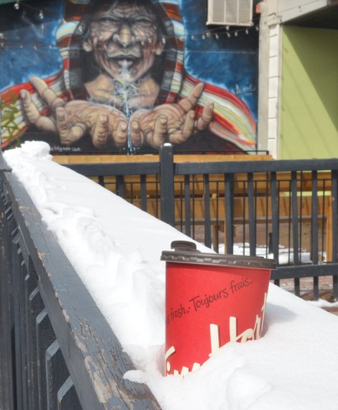 an empty paper coffee cup from Tim Hortons that has been discarded - in the snow on top of a grey wood railing in front of a store. A street art painting of a man with outstretched hands is in the background.