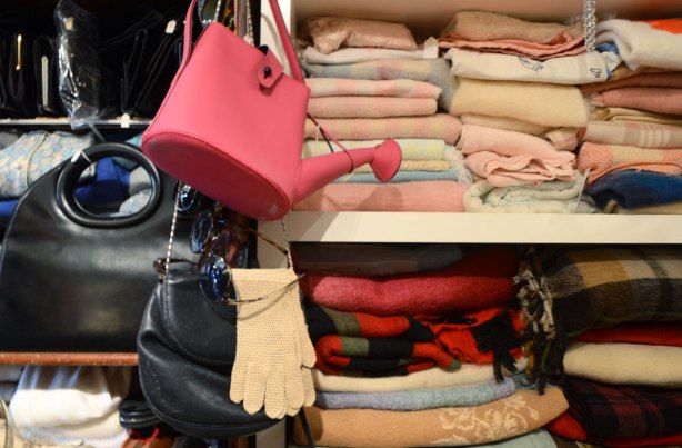 a pink purse in the shape of a watering can hangs from a hook on a wall. A pair of beige gloves hangs below it. Folded fabric items are on the shelves beside the bag and gloves. Blankets or sweaters.