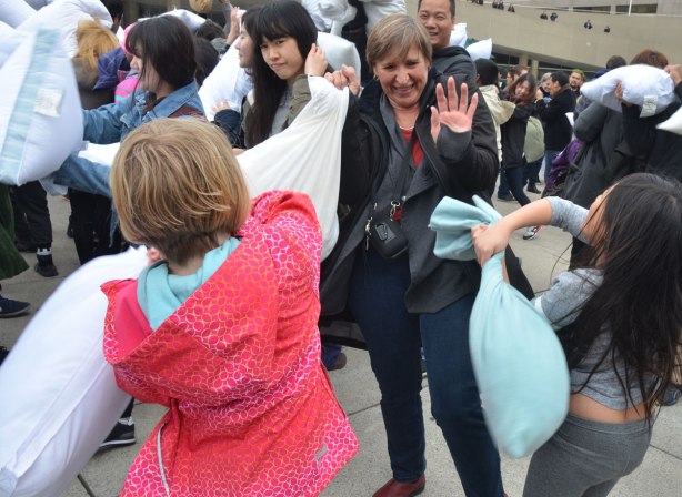 a woman holds her hands up by her head defensively to ward off pillows being swung by two girls
