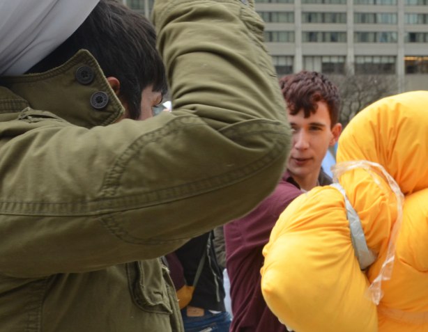 people in the midst of a large pillow fight at Nathan Phillips square in celebration of international pillow fight day - a young man with two yellow pillows looks intensely at a man with a pillow raised over his head
