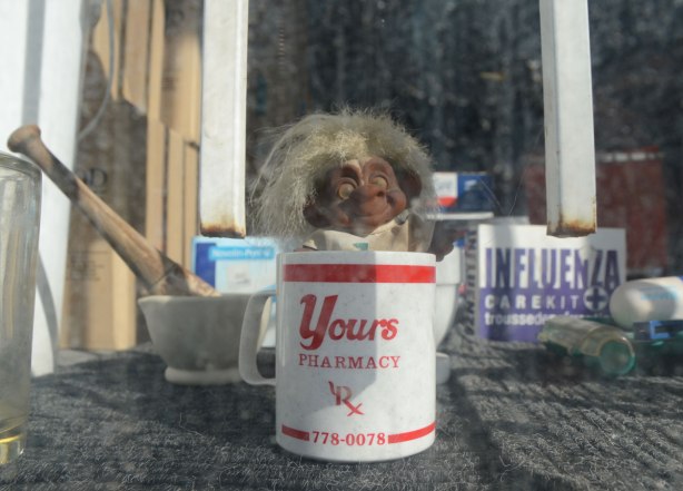 looking into the window of a pharmacy, a toy troll is in a white mug. The mug has red lettering - Yours pharmacy. Also a mortar and pestle in the window along with a box with medicines for influenza