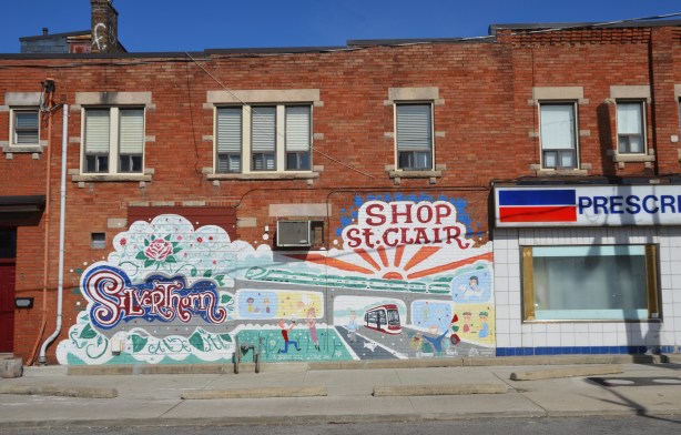 Silverthorn Sunrise mural by the Davenport Arts Community, with the word Silverthorn, a sun rising over a bridge while a new TTC streetcar passes under the bridge, the words "shop St. Clair". The mural is on the side of a red brick building, beside a pharmacy. 