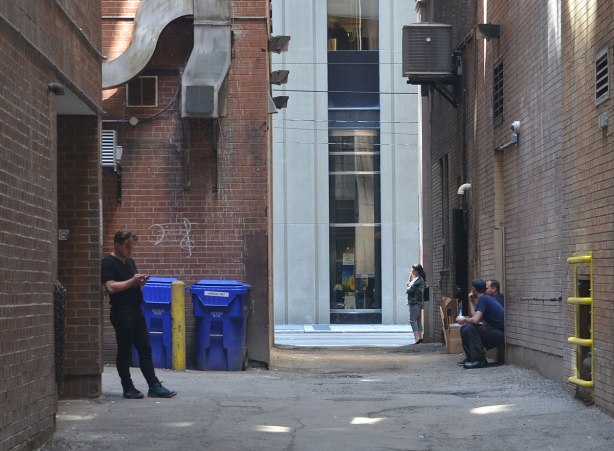 people sitting and standing in an alley, one man is on his cellphone, tall buildings on either side of the lane as well as at the end of the lane 