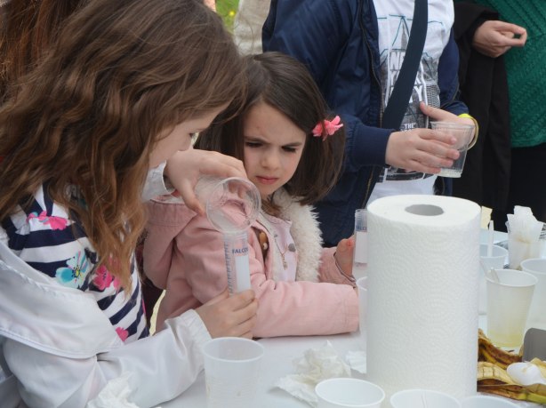 Two young girls are performing an science experiment using beakers and a graduated cylinder. One of them is pouring liquid into the cylinder while the younger one watches. 