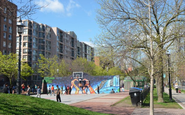 kids playing basketball on an outdoor court. The wall behind the basketball hoop has been painted with a mural of hands making a heart shape with the fingers, by Bruno Smokey and Shalak Attack. 