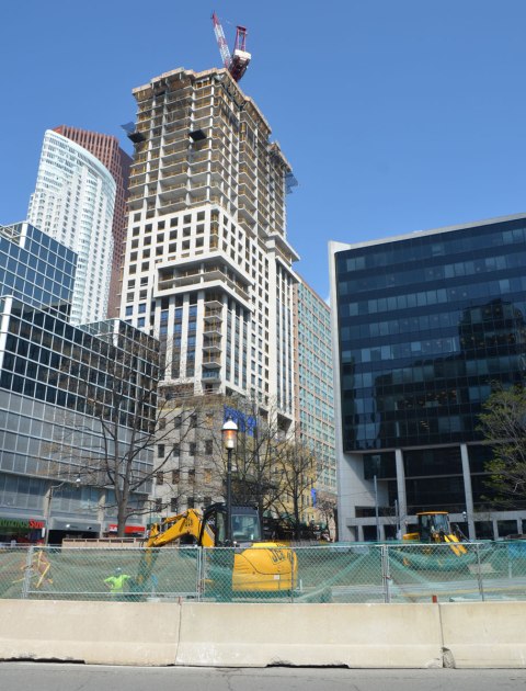 workmen redoing a park, Berczy Park in Toronto, with a digger and another piece of equipment, cityscape behind with a tall condo building under construction. A red and white crane is on top. 