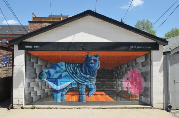 street art mural on a garage door in an alley by birdo - a blue dog beside a pink lava lamp on a table. words: birdo dmc, phil pans tense