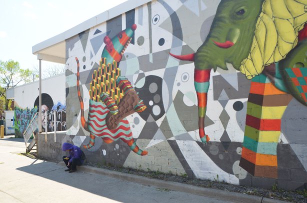 a person is sitting on a kerb in an alley, beside a wall with a large mural by birdo of an elephant and a mouse 