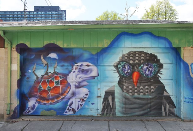 street art mural on a garage door in an alley by birdo - a lavendar turtle and an owl with blue eyes and orange beak